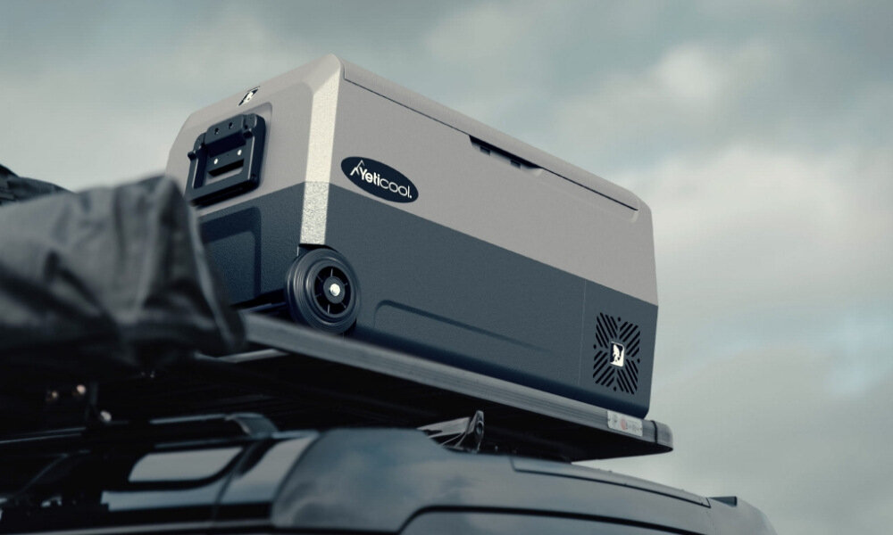 Szara lodówka przenośna z logo \'Yeticool\' umieszczona na dachu pojazdu. W tle widoczne jest pochmurne niebo.