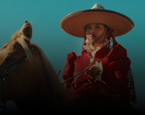 Osoba w czerwonym stroju z dużym kapeluszem siedzi na koniu. Tło jest jasnoniebieskie.