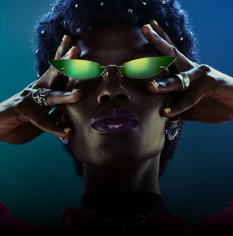Osoba nosi zielone okulary przeciwsłoneczne o nietypowym kształcie, z dłońmi uniesionymi do twarzy. Tło jest ciemne z niebiesko-zielonym gradientem.