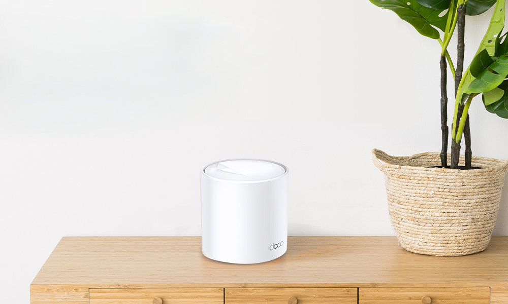 Na drewnianej powierzchni stoi biały, cylindryczny router z napisem \'deco\'. Obok routera znajduje się roślina w plecionym koszu.