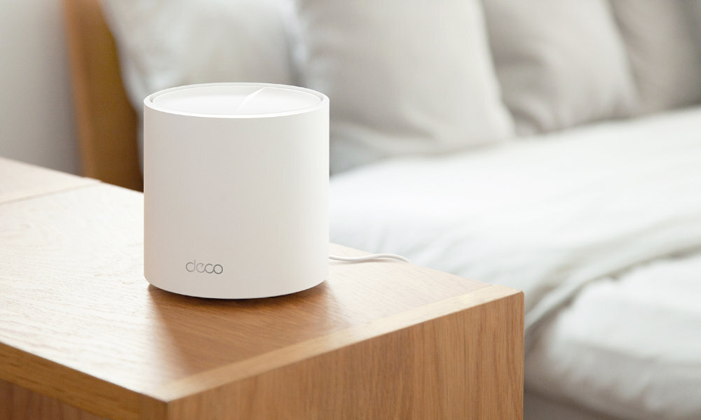 Na drewnianym stoliku nocnym stoi biały, cylindryczny router z napisem \'deco\'. W tle widoczne jest łóżko z białą pościelą.