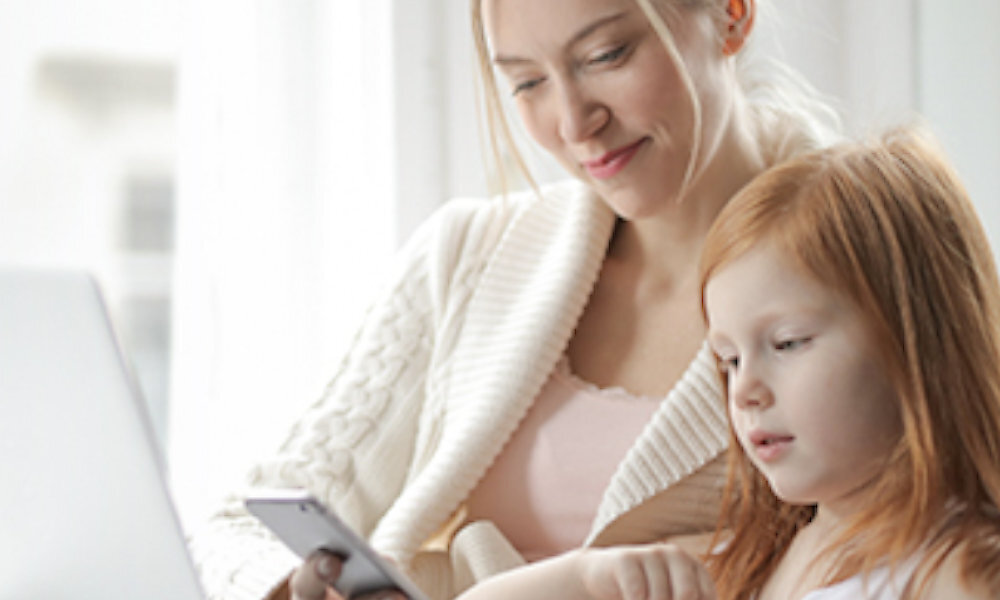 Na obrazie widać dwie osoby, dorosłą i dziecko, siedzące razem. Dorosła osoba trzyma smartfon, a dziecko patrzy na ekran laptopa.