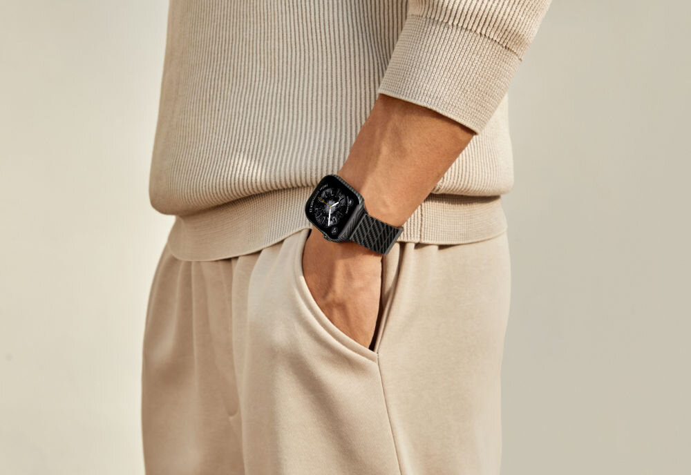 Osoba ubrana w beżowy sweter i spodnie ma na nadgarstku czarny zegarek z ciemnym paskiem. Zegarek ma wyświetlacz z widocznym tarczą.