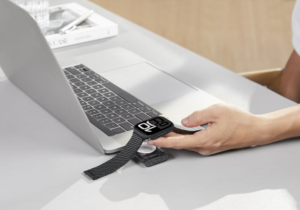 Osoba trzyma czarno-szary zegarek z wyświetlaczem pokazującym godzinę 21:28, który jest podłączony do ładowarki obok laptopa. Na stole leży książka z napisem \'CASE\'.