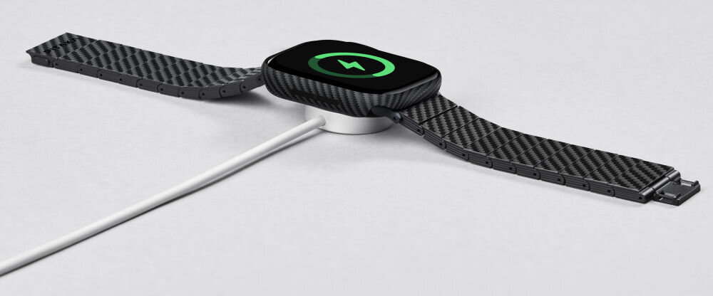 Na obrazie widać zegarek z czarno-szarym paskiem o wzorze przypominającym włókno węglowe, umieszczony na ładowarce bezprzewodowej. Na ekranie zegarka widoczna jest ikona ładowania z zielonym symbolem błyskawicy.