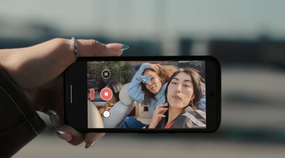 Osoba trzyma smartfon, na ekranie którego widać aplikację do nagrywania wideo z dwiema osobami w kadrze. W tle rozmyte elementy krajobrazu.