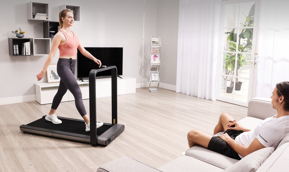 W jasnym salonie kobieta ćwiczy na bieżni elektrycznej, podczas gdy mężczyzna siedzi na kanapie. W tle widać telewizor, półki z książkami i rośliny przy oknie.