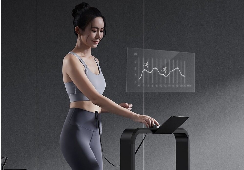 Osoba stoi przy bieżni elektrycznej, patrząc na ekran z wykresem przedstawiającym biegaczy i linie trendu. Bieżnia ma nowoczesny design z zakrzywionym uchwytem i panelem sterowania.