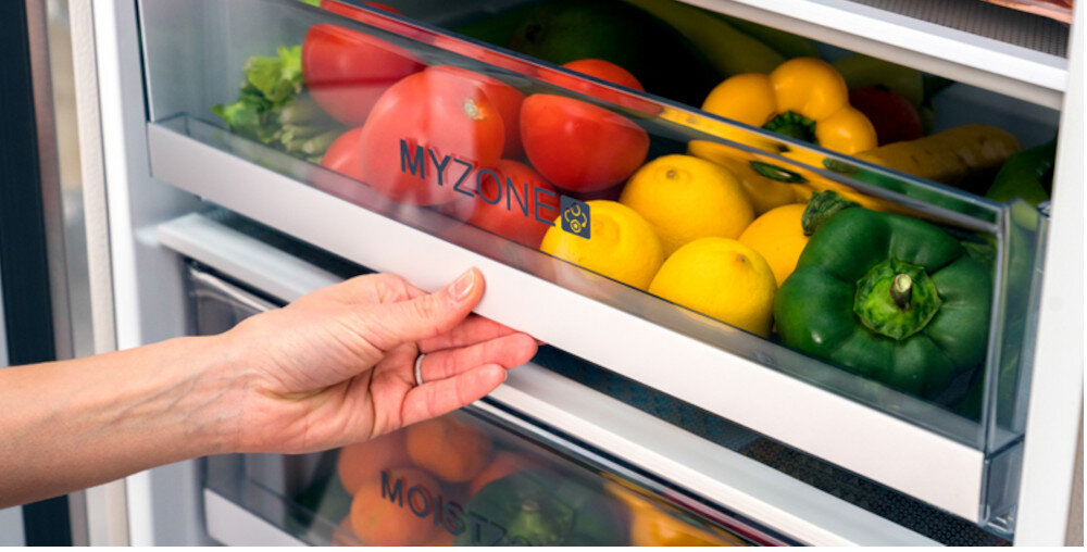 Osoba otwiera szufladę lodówki z napisem \'MYZONE\', w której znajdują się różne warzywa i owoce, takie jak pomidory, cytryny i papryki.