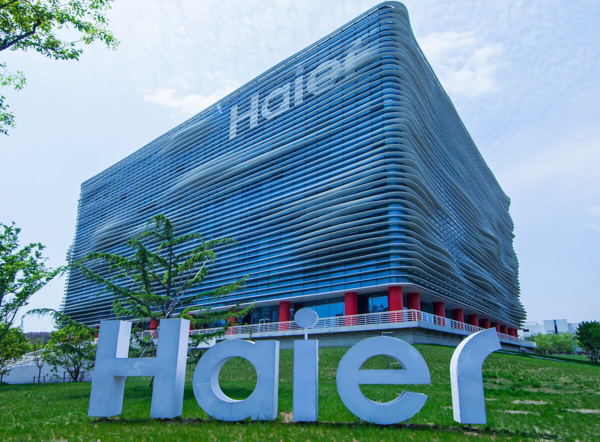 Duży, nowoczesny budynek z falistą fasadą i napisem \'Haier\' na górze. Przed budynkiem znajduje się duże logo \'Haier\' na trawniku.