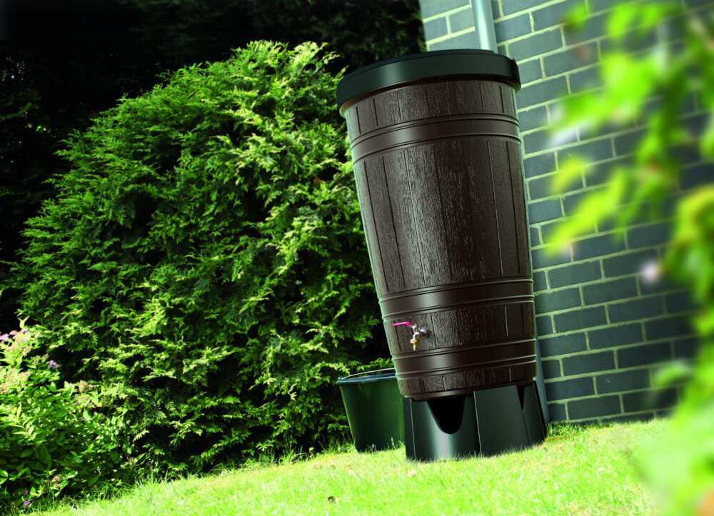 Na obrazie widoczny jest brązowy zbiornik na deszczówkę o wyglądzie przypominającym drewnianą beczkę, ustawiony na trawie obok ceglanego muru. W tle znajduje się zielony krzew.