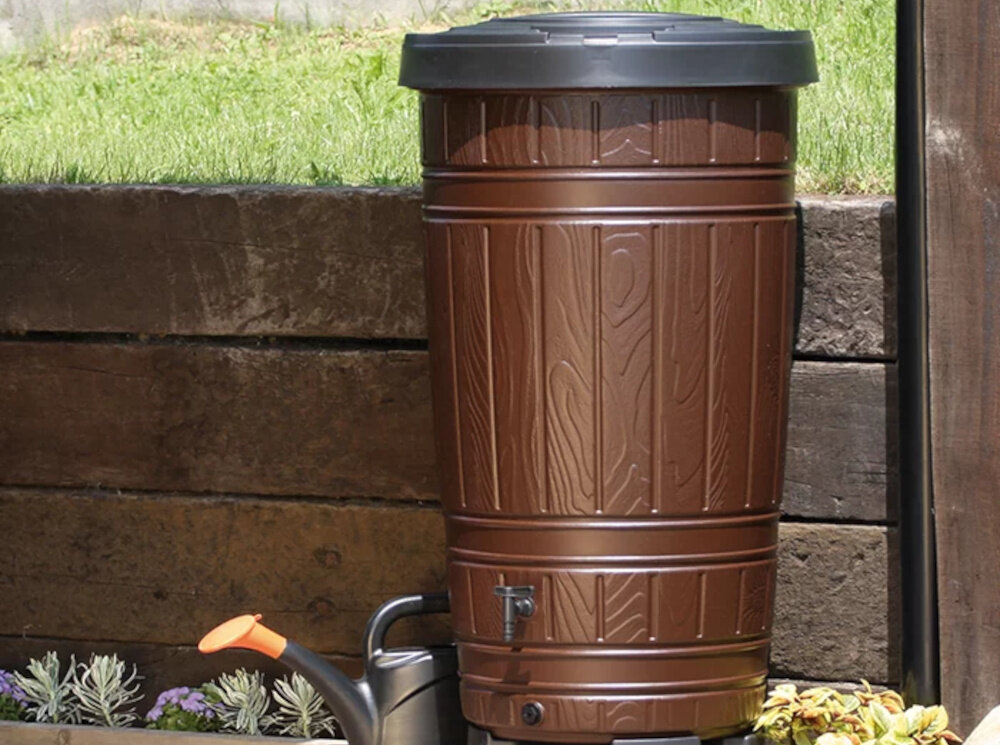 Brązowy zbiornik na deszczówkę o fakturze przypominającej drewno, z czarną pokrywą na górze. Obok znajduje się konewka z pomarańczowym uchwytem.