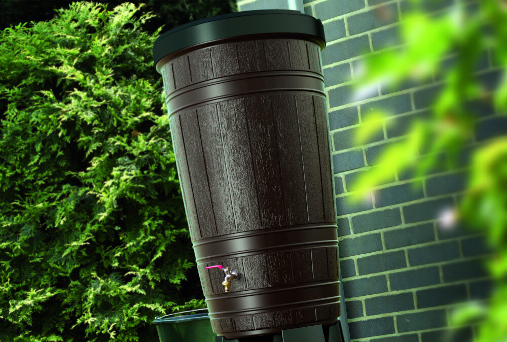 Brązowy zbiornik na deszczówkę o wyglądzie drewnianej beczki, z kranikiem na dole, stoi obok ceglanej ściany. W tle widoczne są zielone krzewy.