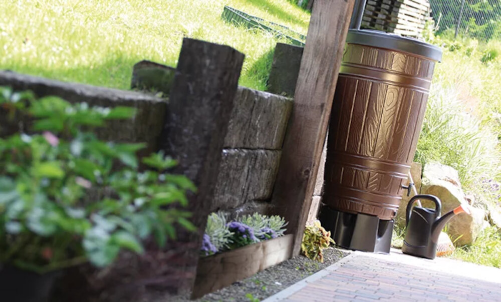 Brązowy zbiornik na deszczówkę o fakturze przypominającej drewno stoi obok drewnianego płotu. Obok znajduje się czarna konewka na tle zielonej trawy i kamieni.