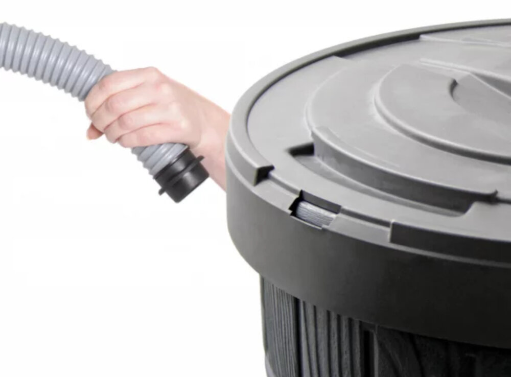 Osoba trzyma szary, elastyczny wąż z czarną końcówką. Obok znajduje się górna część brązowego zbiornika na deszczówkę z pokrywą.