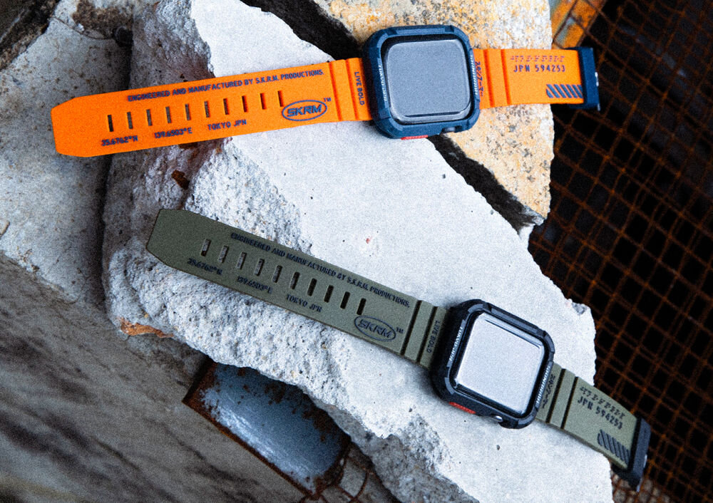 Na zdjęciu widoczne są dwa paski do zegarków Apple Watch, jeden w kolorze oliwkowym, drugi pomarańczowy, leżące na betonowych blokach. Na obu paskach znajduje się tekst: \'ENGINEERED AND MANUFACTURED BY S.K.R.M. PRODUCTIONS\' oraz logo \'SKRM\'.