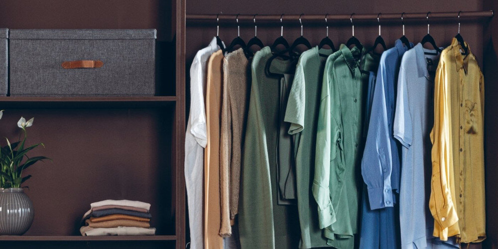 Szafa z ubraniami na wieszakach, w różnych kolorach, obok półki z szarą pudełkiem i złożonymi ubraniami. Na dolnej półce stoi wazon z rośliną.