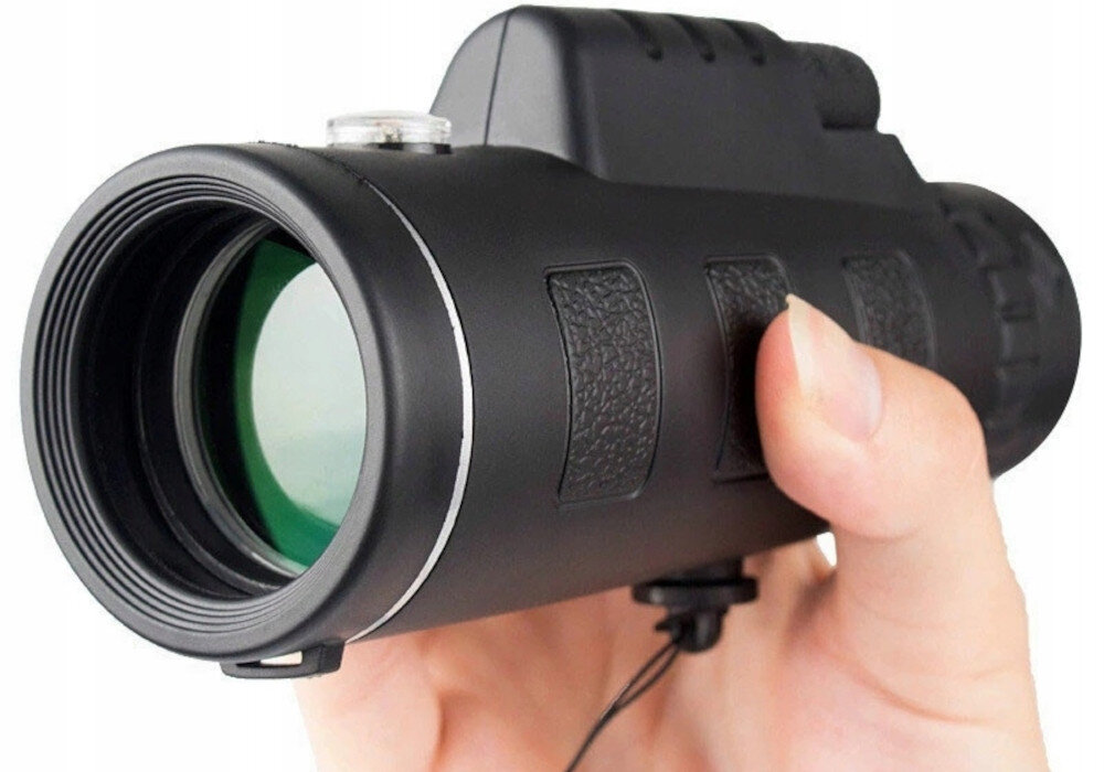 Osoba trzyma czarny monokular z zielonym obiektywem i teksturowanymi uchwytami. Monokular ma cylindryczny kształt i jest wyposażony w pasek na nadgarstek.