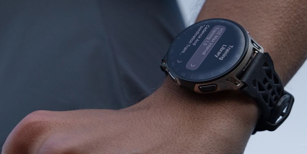 Osoba nosi smartwatch z okrągłym wyświetlaczem, na którym widoczny jest tekst: \'Training Library\'. Pasek zegarka ma ażurowy wzór.
