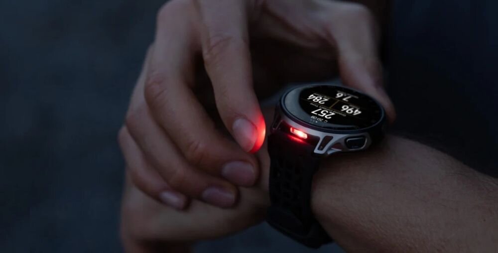 Osoba nosi smartwatch z okrągłym wyświetlaczem, na którym widoczne są cyfry i dane. Zegarek emituje czerwone światło z boku obudowy.