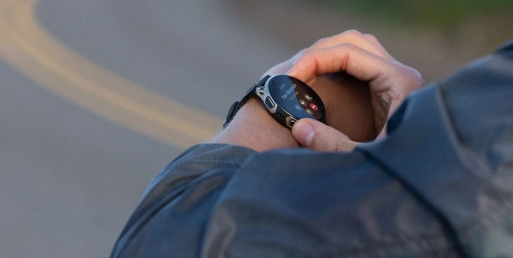 Osoba nosi smartwatch na nadgarstku, ekran urządzenia jest włączony i wyświetla różne informacje. W tle widoczna jest rozmyta droga.