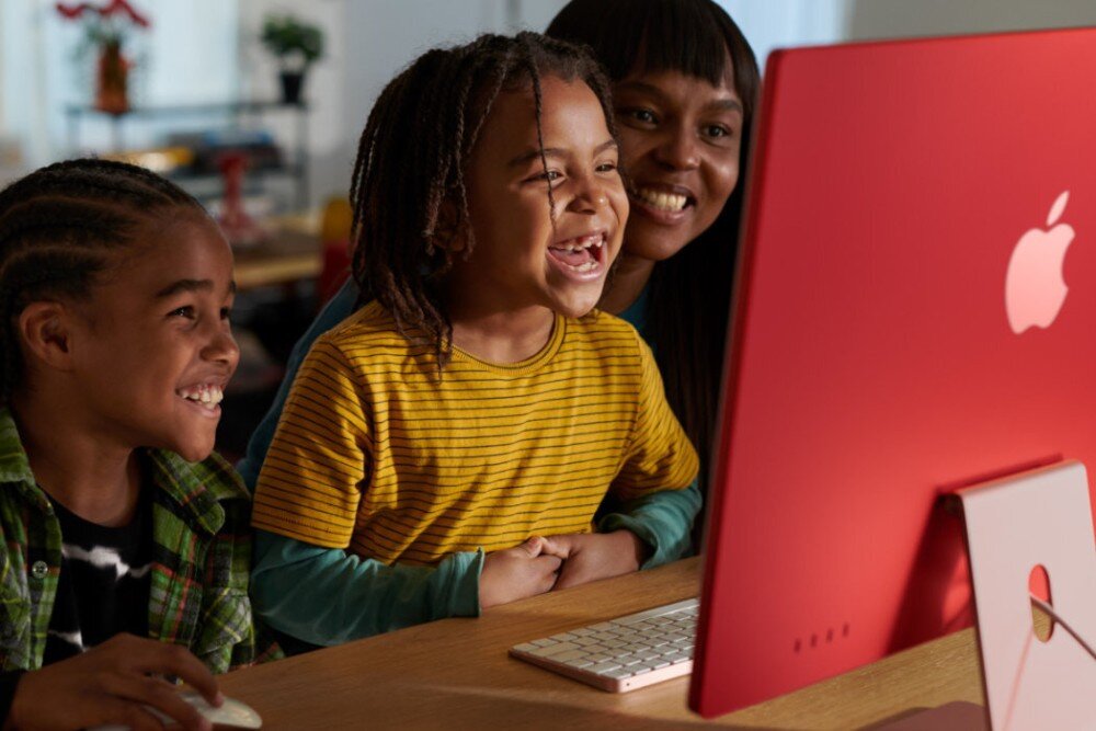 Na obrazie widoczny jest różowy komputer All in One z logo w kształcie jabłka. Przed komputerem siedzą trzy osoby, które patrzą na ekran.