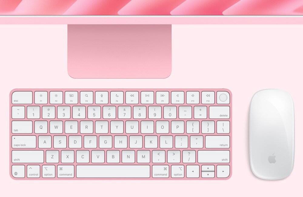 Na obrazie widoczna jest różowa klawiatura z białymi klawiszami oraz biała myszka z logo w kształcie jabłka. W tle znajduje się różowa podstawa monitora.