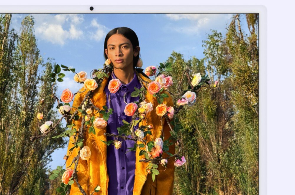 Osoba stoi na tle drzew, ubrana w żółty płaszcz i fioletową koszulę, ozdobiona gałązkami z różowymi i pomarańczowymi kwiatami. W tle widać niebo z białymi chmurami.