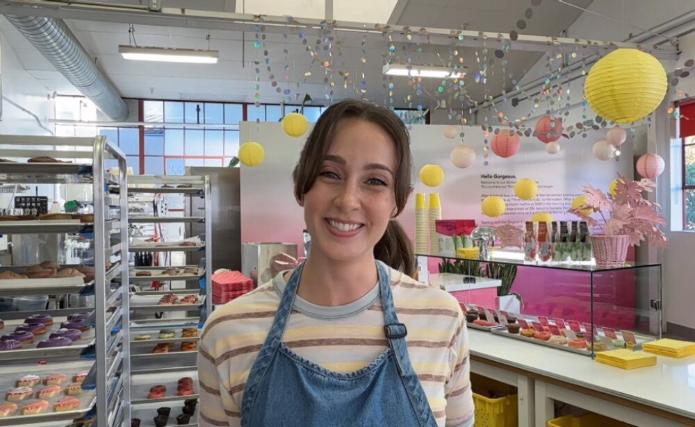 Wnętrze sklepu z wypiekami, z dekoracjami w postaci kolorowych kul i girland. Na pierwszym planie osoba w fartuchu stoi przed ladą z ciastkami i napojami.