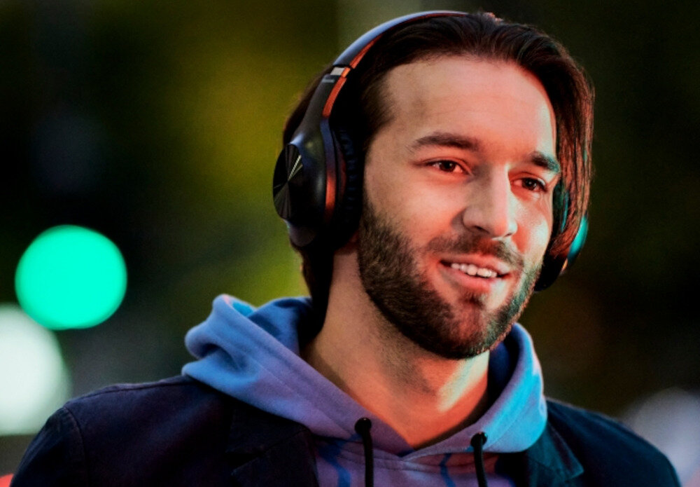 Osoba nosi słuchawki nauszne w ciemnym kolorze, ubrana w niebieską bluzę z kapturem. Tło jest rozmyte z widocznymi kolorowymi światłami.