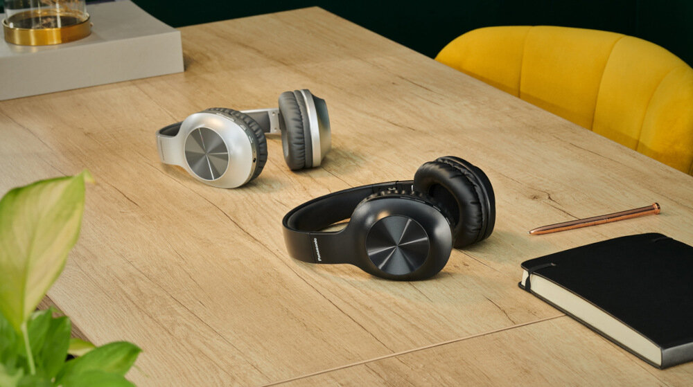 Na drewnianym stole leżą dwie pary słuchawek nausznych, jedna srebrna, druga czarna. Obok znajduje się żółte krzesło, notes i długopis.