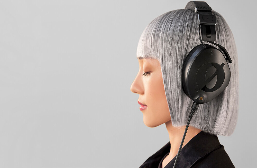 Osoba z krótkimi, srebrnymi włosami nosi czarne słuchawki nauszne. Tło jest jednolite i jasne.