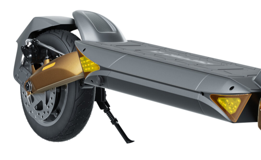 Zdjęcie przedstawia tylną część hulajnogi elektrycznej z widocznym kołem, amortyzatorem i podnóżkiem. Elementy są w kolorze szarym i miedzianym, z żółtymi odblaskami.