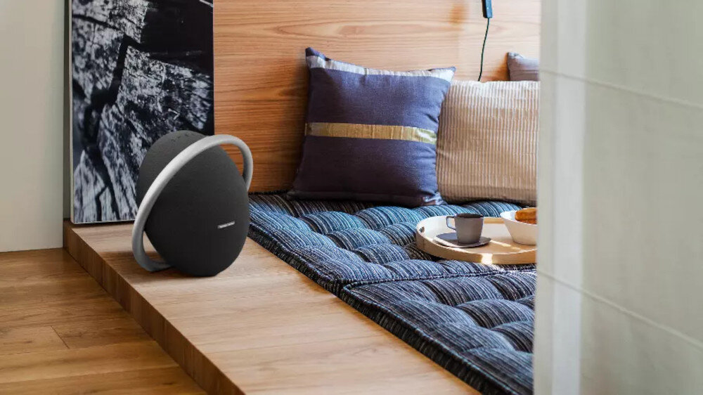 Na drewnianej podłodze znajduje się czarny głośnik o okrągłym kształcie z metalicznym uchwytem. Obok głośnika leży mata z poduszkami oraz taca z filiżanką i talerzykiem.