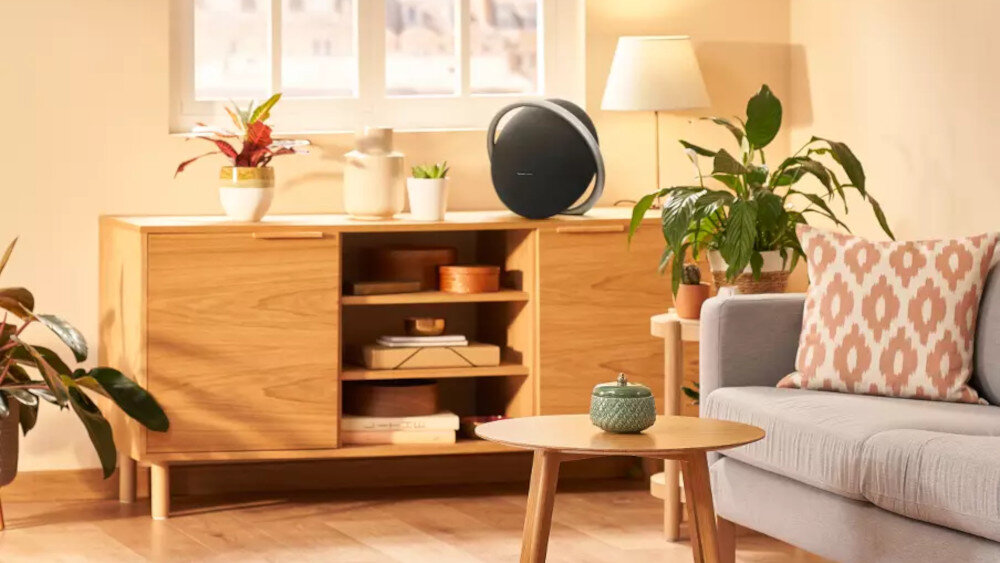 W salonie na drewnianej szafce stoi czarny głośnik o okrągłym kształcie. W pomieszczeniu znajdują się również rośliny, lampa i sofa z poduszką w geometryczny wzór.