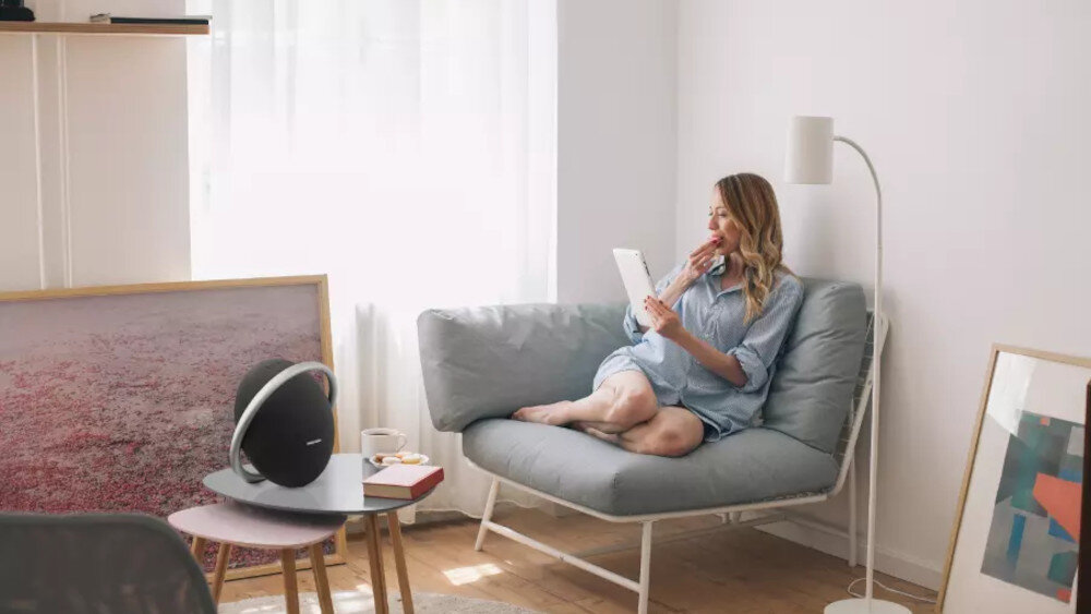 W jasnym pokoju na stoliku znajduje się czarny głośnik mobilny z widocznym logo \'Harman Kardon\'. Obok stolika siedzi osoba na sofie, trzymająca tablet.