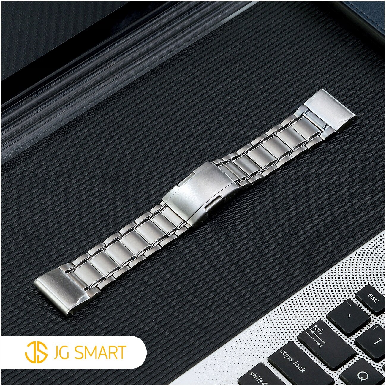 Na obrazie widoczna jest metalowa bransoleta zegarka umieszczona na ciemnej, prążkowanej powierzchni obok klawiatury laptopa. W lewym dolnym rogu znajduje się logo \'JG SMART\'.