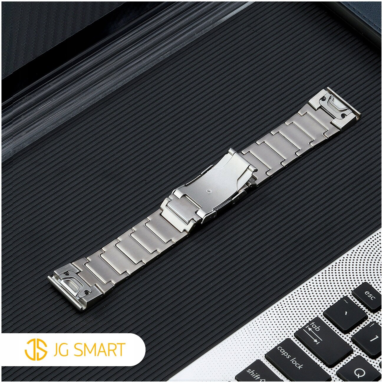Na obrazie widoczna jest metalowa bransoleta na ciemnym tle, obok fragmentu klawiatury. W lewym dolnym rogu znajduje się logo \'JG SMART\'.