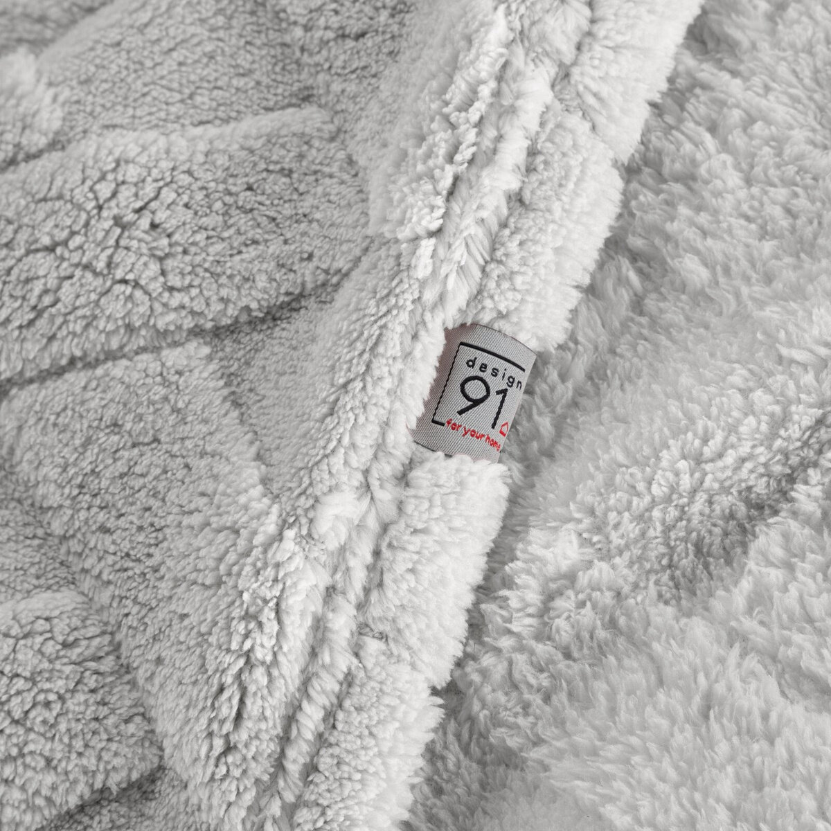Na obrazie widoczny jest srebrny, pluszowy koc z wzorem jodełki. Na kocu znajduje się metka z napisem \'design 91 for your home\'.