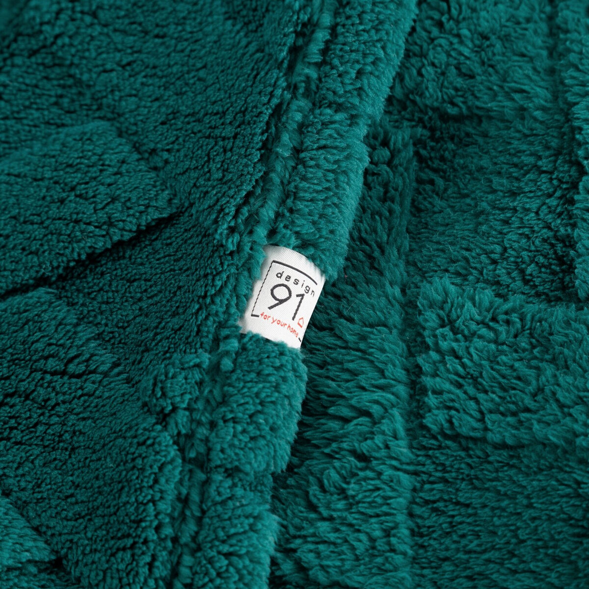 Pluszowy koc w kolorze ciemnej zieleni z widoczną metką z napisem \'design 91 for your home\'.