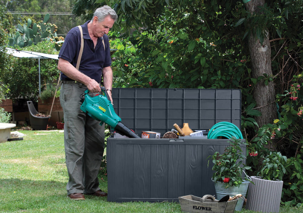 Osoba stoi przy otwartej skrzyni ogrodowej, w której znajdują się różne narzędzia i przedmioty ogrodowe. W tle widoczna jest zieleń ogrodu.