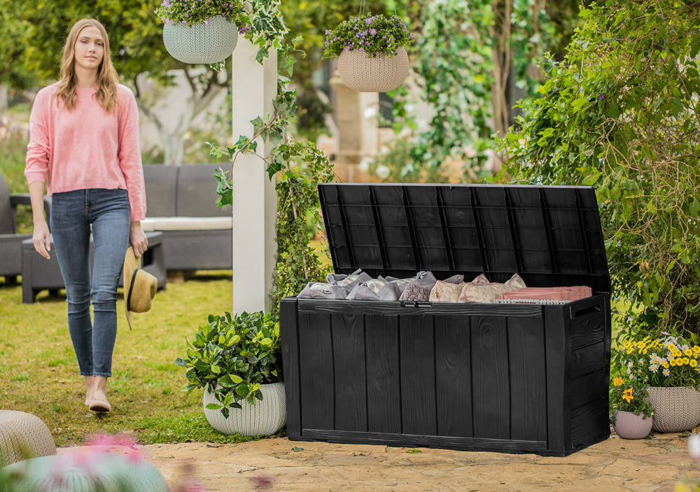Otwarta skrzynia ogrodowa w kolorze brązowym, wypełniona poduszkami, stoi na trawniku w otoczeniu roślin. W tle widoczna jest osoba oraz meble ogrodowe.