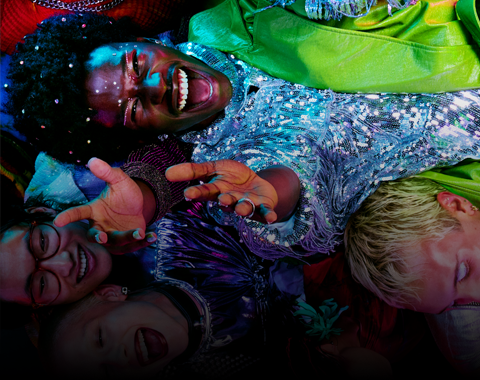 Grupa osób leży blisko siebie, uśmiechając się i patrząc w górę. Osoba w centrum ma na sobie błyszczącą, cekinową bluzkę.