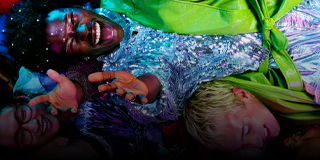 Grupa osób ubranych w kolorowe, błyszczące stroje leży blisko siebie, jedna z nich szeroko się uśmiecha.