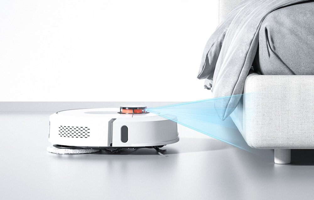 Biały robot sprzątający z czarnymi i czerwonymi elementami porusza się obok łóżka, emitując niebieski promień światła w kierunku mebla. Podłoga jest jasna, a łóżko pokryte jest szarą pościelą.