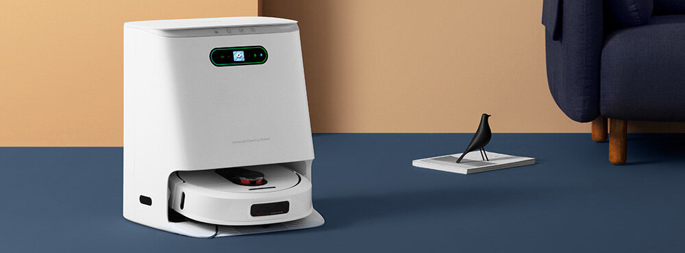 Na obrazie widoczny jest biały robot sprzątający w stacji dokującej na niebieskiej podłodze, obok stoi czarna figurka ptaka na książce. W tle znajduje się fragment ciemnej sofy na tle beżowej ściany.