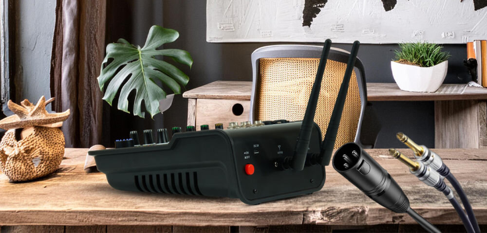 Na drewnianym biurku znajduje się mikser audio z widocznymi pokrętłami i dwoma antenami. Obok leżą kable z wtykami XLR i jack.