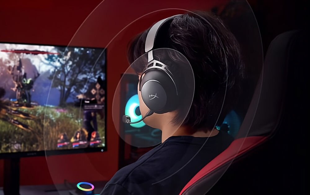 Osoba siedzi na fotelu przed monitorem, nosząc nauszne słuchawki z mikrofonem. Na ekranie widoczna jest gra komputerowa z postaciami i krajobrazem.