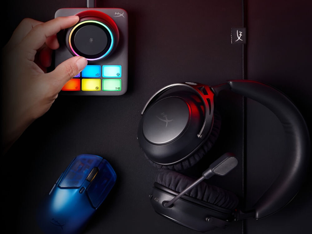 Na obrazie widoczne są czarne słuchawki nauszne z mikrofonem oraz ręka obsługująca urządzenie z kolorowymi przyciskami i pokrętłem. Obok znajduje się przezroczysta, niebieska myszka komputerowa.