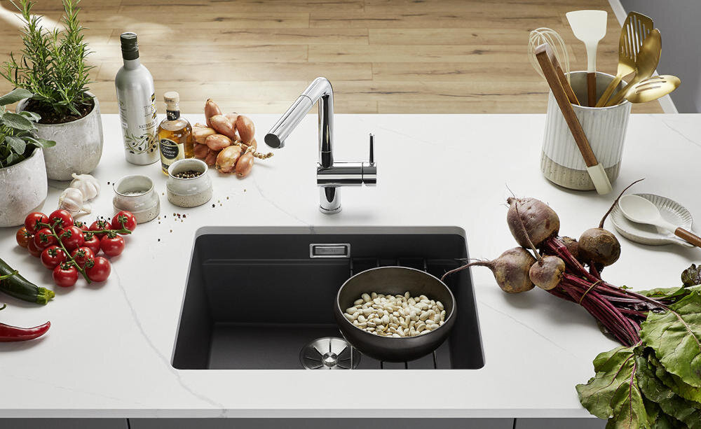 Czarny zlewozmywak jednokomorowy z zamontowaną baterią, w którym znajduje się miska z białymi fasolami. Na blacie wokół zlewozmywaka znajdują się różne warzywa, przyprawy i akcesoria kuchenne.